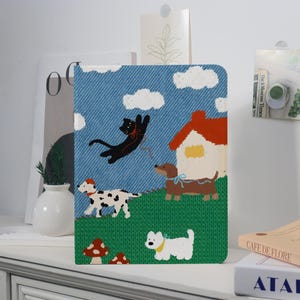 May include: A decorative notebook with a whimsical illustration. The cover features a blue denim-like background with a cartoon scene of a black cat, dogs, a house, clouds, and mushrooms. The notebook is standing upright on a white surface.