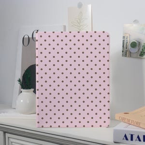 Könnte beinhalten: Ein rosa Notizbuch mit einem braunen Polka-Dot-Muster. Das Notizbuch steht auf einer weißen Oberfläche. Das Cover ist glatt und die Punkte sind gleichmäßig verteilt.