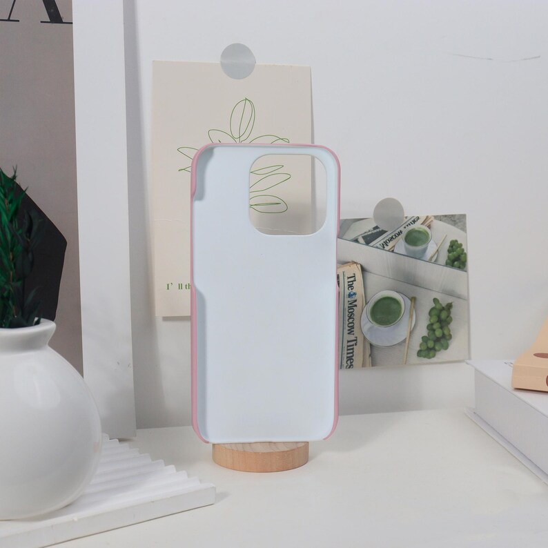 K&ouml;nnte beinhalten: Eine wei&szlig;e Handyh&uuml;lle mit rosa Rand, die auf einem kleinen Holzst&auml;nder steht. Die H&uuml;lle ist leer, mit Aussparungen f&uuml;r Kamera und Kn&ouml;pfe. Der Hintergrund umfasst dekorative Elemente und einen Zeitungsausschnitt.
