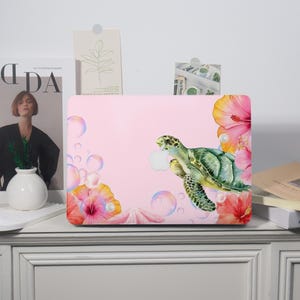 May include: A pink laptop case featuring a watercolor design of a green sea turtle holding an apple. The case is decorated with hibiscus flowers, bubbles, and a seashell. The design has a tropical theme.