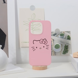 May include: A pink phone case featuring a Hello Kitty design. The case has a cutout for the camera and buttons. The Hello Kitty face is drawn in black, with whiskers and a bow. The case is standing on a small wooden stand.