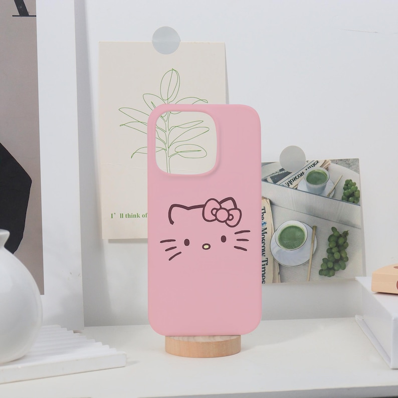 K&ouml;nnte beinhalten: Eine rosa Handyh&uuml;lle mit einem Hello Kitty-Design. Die H&uuml;lle hat Aussparungen f&uuml;r Kamera und Kn&ouml;pfe. Das Hello Kitty-Gesicht ist in Schwarz gezeichnet, mit Schnurrhaaren und einer Schleife. Die H&uuml;lle steht auf einem kleinen Holzst&auml;nder.