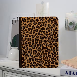 May include: A notebook with a leopard print cover. The cover features a pattern of brown and black spots on a tan background. The notebook is closed and has a dark brown spine.