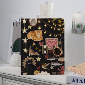 May include: A black notebook featuring a collage of cat illustrations, stars, hearts, and other decorative elements. The cover includes a variety of cat breeds in different poses, with a string of lights and star accents.