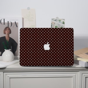 May include: A dark brown laptop case with a white polka dot pattern. The Apple logo is visible in the center. The case is on a white surface with decorative items in the background.