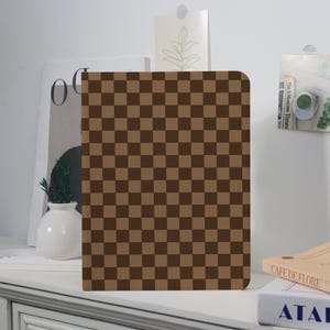 May include: A brown and tan chequered pattern notebook or tablet cover. The cover is rectangular with rounded corners. The pattern is a repeating grid of dark brown and tan squares. The cover is standing upright on a white surface.