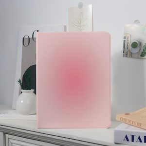 Könnte beinhalten: Eine rosa Tablet-Hülle mit Farbverlauf, von dunklerem Rosa in der Mitte zu hellerem Rosa an den Rändern. Die Hülle steht auf einer weißen Oberfläche, mit anderen dekorativen Gegenständen im Hintergrund.