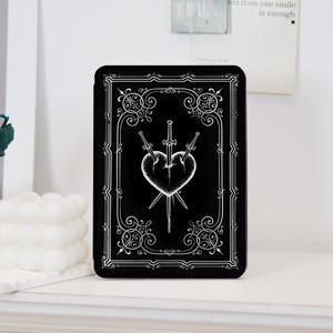 May include: A black e-reader case featuring a white line art illustration of a heart pierced by three swords. The design is enclosed within an ornate, rectangular border with decorative scrollwork at each corner, mimicking a playing card aesthetic.
