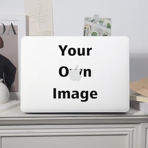 May include: A white laptop with the words "Your Own Image" in black text. The Apple logo is visible. The laptop is on a white surface with magazines and decorative items in the background.