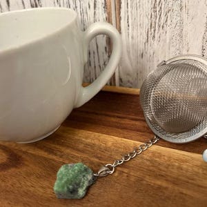 May include: A white ceramic teacup with a curved handle sits next to a stainless steel tea infuser. The infuser has a chain attached to a green and black stone. The items are on a wooden tray.