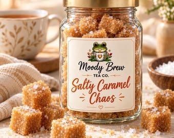 Puede incluir: Un frasco de vidrio transparente lleno de cubos de azúcar moreno, con una tapa dorada. La etiqueta muestra una ilustración de una rana verde y el texto Moody Brew Tea Co. Salty Caramel Chaos. Varios cubos de azúcar están esparcidos.