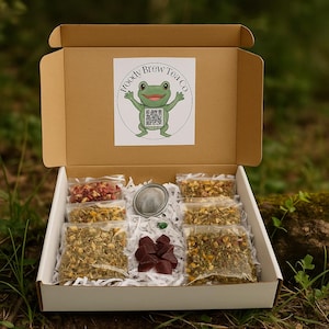 May include: An open cardboard box filled with tea and tea accessories. The box features a logo with a green frog. Inside are several small bags of tea, a tea infuser, and some red cubes. The box is on a white surface.