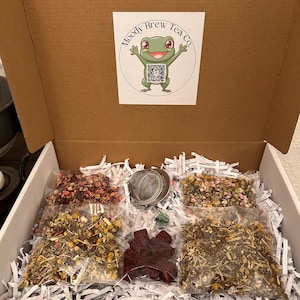 May include: A cardboard box filled with tea and accessories. The box contains several clear plastic bags of loose leaf tea, a metal tea infuser, and a decorative piece. The box has a sticker with a frog logo and the text "Moody Brew Tea Co."