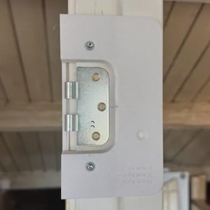 Peut inclure: Un cache-charnière de porte blanc avec une charnière métallique et des vis. Le cache est rectangulaire avec des coins arrondis et est monté sur un cadre de porte blanc. La charnière est argentée et comporte trois trous de vis visibles.