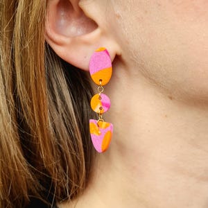 Puede incluir: Pendientes colgantes con un diseño abstracto y vibrante. Los pendientes presentan una combinación de colores rosa y naranja en un patrón geométrico en capas. Los pendientes están conectados por pequeños aros dorados.