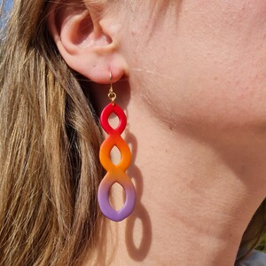 Pode incluir: Um par de brincos longos e coloridos com um design gradiente. Os brincos fazem uma transição de vermelho para laranja e roxo. Os brincos estão pendurados em um gancho dourado.