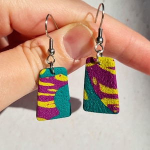 Może przedstawiać: Para prostokątnych kolczyków wiszących. Kolczyki mają turkusową podstawę z wzorem żółtych pasków i fioletowych kształtów. Zawieszone są na srebrnych haczykach.