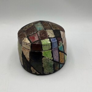 May include: A decorative, handcrafted box with a lid. The box is covered in a mosaic pattern of colorful squares in shades of brown, green, red, and blue. The box has a rounded shape and appears to be made of a textured material.