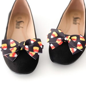 Puede incluir: Un par de bailarinas negras con lazos con estampado de maíz dulce. Los lazos están atados en un nudo en la parte superior de los zapatos.