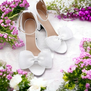 May include: A pair of white satin bridal heels with delicate white bows. The heels are on a white background with pink flowers.