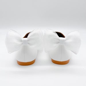 May include: A pair of white leather ballet flats with a large white bow on each shoe. The shoes have a brown sole.