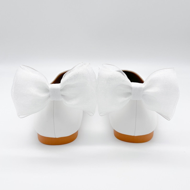 May include: A pair of white leather ballet flats with a large white bow on each shoe. The shoes have a brown sole.