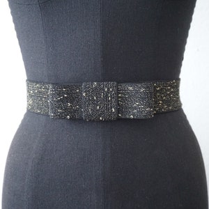 May include: A black and gold elastic belt with a bow in the centre. The belt is made of a woven fabric with gold thread woven throughout.