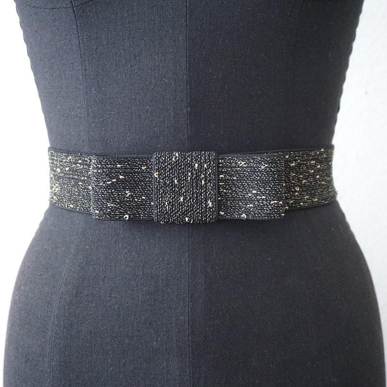 May include: A black and gold elastic belt with a bow in the centre. The belt is made of a woven fabric with gold thread woven throughout.