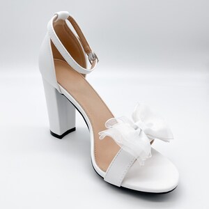 May include: White high-heeled sandals with a delicate white bow on the front strap. The heels are block shaped and the sandals have a closed toe.