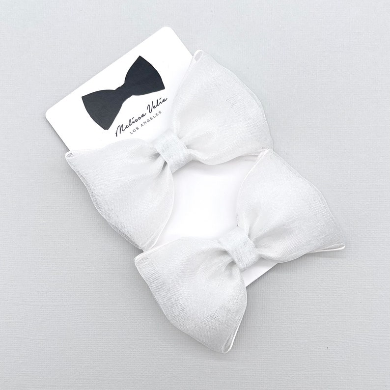 May include: Two white satin ribbon bows with a white card behind them. The card has the text "Melissa Vale, Los Angeles" printed on it.