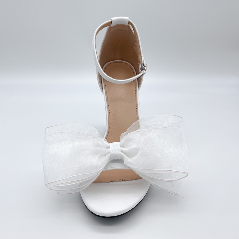 May include: White high-heeled sandals with a large white bow made of tulle.