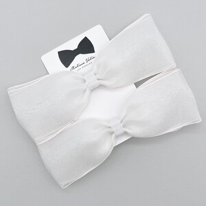 May include: Two white sheer ribbon bows with a white satin center. The bows are on a white card with the text "Melissa Vida Los Angeles" and a black bow tie.