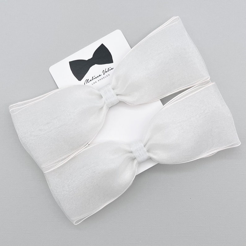 May include: Two white sheer ribbon bows with a white satin center. The bows are on a white card with the text "Melissa Vida Los Angeles" and a black bow tie.