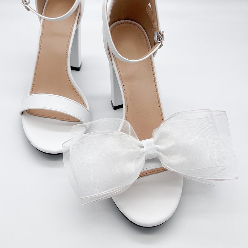 May include: White high-heeled sandals with a large white bow on the front.