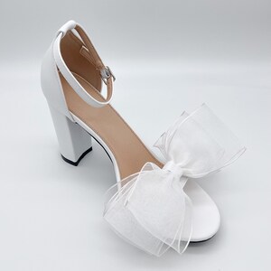 May include: White high-heeled sandals with a large white bow on the front. The sandals have a thick block heel and an ankle strap.