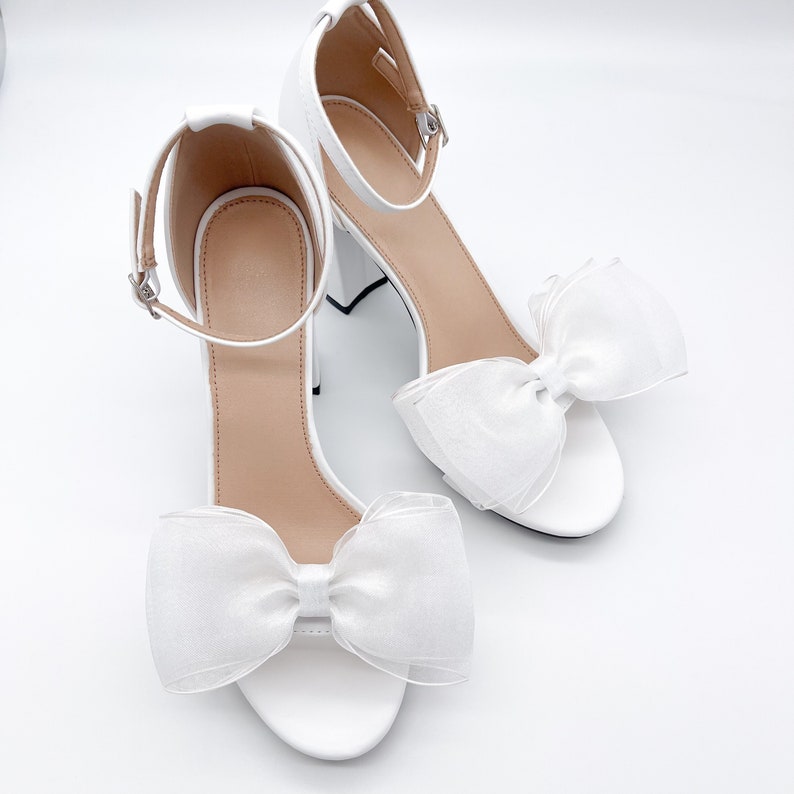 May include: White bridal heels with a large white bow on each shoe. The heels are ankle strap sandals with a closed toe.