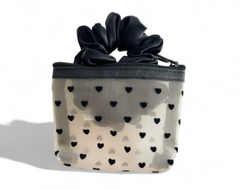 May include: A black mesh pouch with a zipper closure and a black satin scrunchie on top. The pouch has a white background with black heart-shaped patterns.