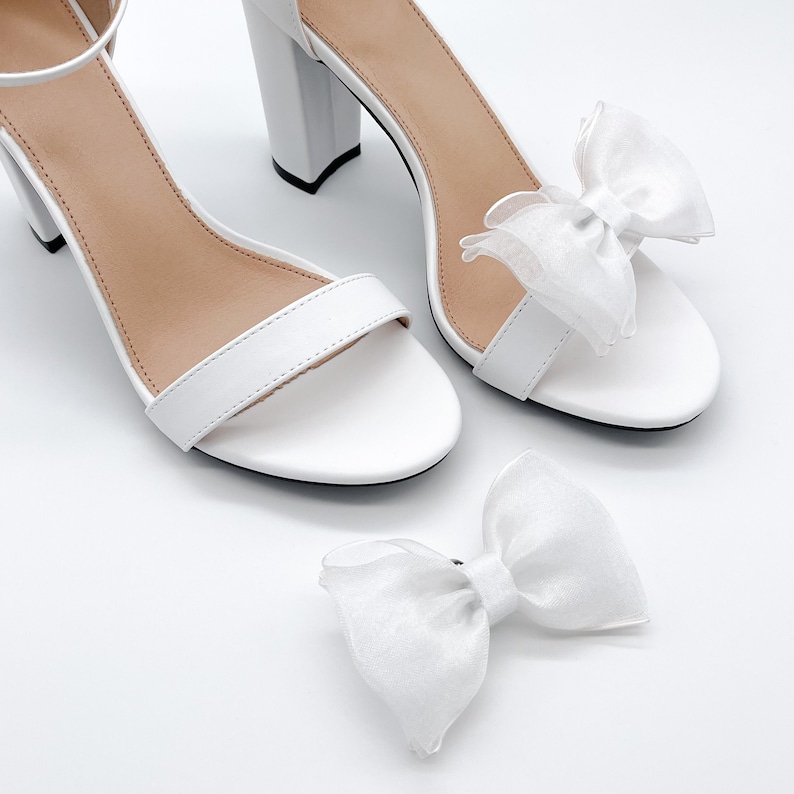May include: A pair of white heeled sandals with a delicate white bow on each strap. The sandals have a simple design and are perfect for a wedding or other special occasion.
