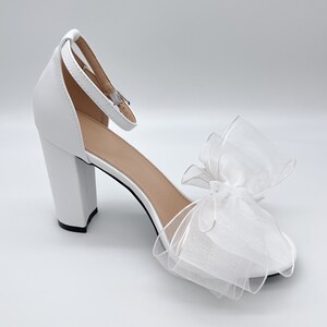 May include: White high-heeled sandals with a large white bow on the toe. The sandals have a thick block heel and an ankle strap.
