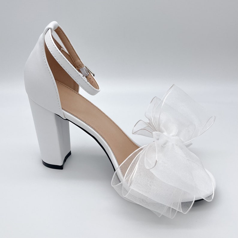 May include: White high-heeled sandals with a large white bow on the toe. The sandals have a thick block heel and an ankle strap.
