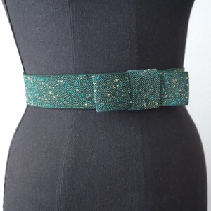 May include: A green and gold sparkly belt with a bow. The belt is made of a woven fabric with a subtle shimmer.