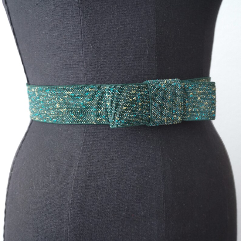 May include: A green and gold sparkly belt with a bow. The belt is made of a woven fabric with a subtle shimmer.