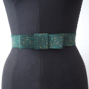 May include: A dark green belt with a gold and teal speckled pattern. The belt has a large bow at the front.