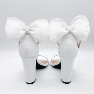 May include: White high-heeled sandals with a large white bow on each ankle strap.