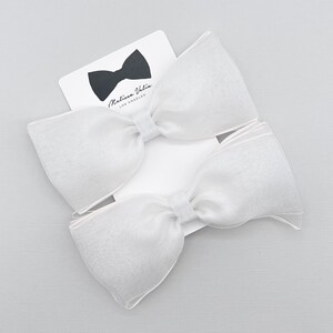 May include: Two white sheer fabric bow hair clips. The bows are large and have a slightly ruffled edge. The bows are on a white background with a small white card with the text "Malina Vela Los Angeles" in black.