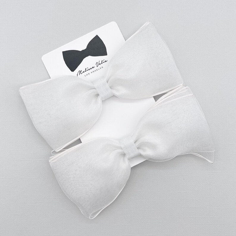 May include: Two white sheer fabric bow hair clips. The bows are large and have a slightly ruffled edge. The bows are on a white background with a small white card with the text "Malina Vela Los Angeles" in black.
