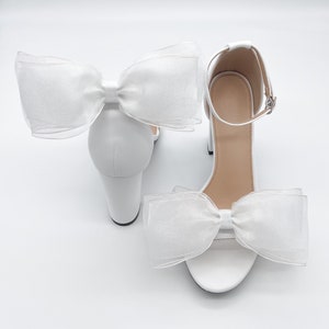 May include: White bridal heels with a large white bow on each shoe. The heels are made of a satin-like material and have a block heel.