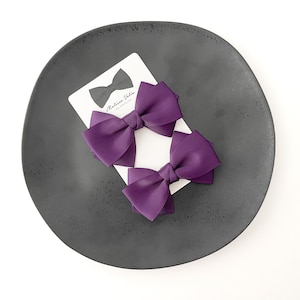 May include: Two purple satin hair bows on a black plate with a white card that says "Madeline Ulloa Los Angeles".