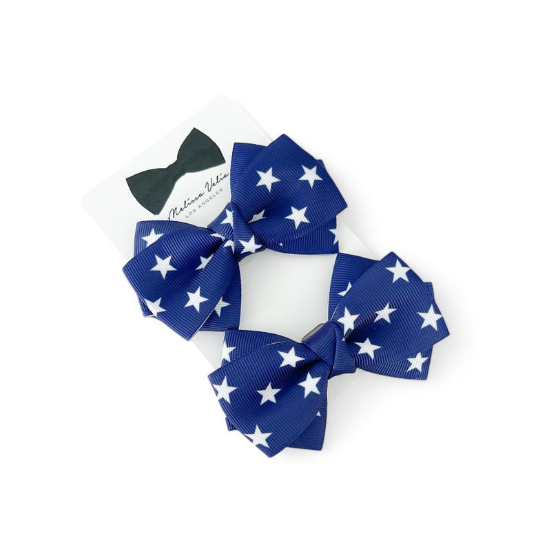 May include: Two navy blue hair bows with white stars. The bows are made of grosgrain ribbon and have a clip on the back.