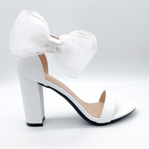 May include: White high-heeled sandals with a large white bow on the ankle strap.